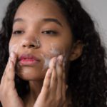 Girl, Wash Your Face - African American female with long dark curly hair washing cheeks with facial foam on gray background